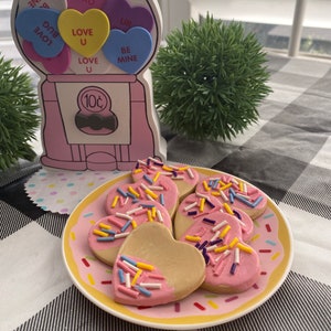 May include: A plate of pink heart-shaped cookies with colorful sprinkles. The cookies are arranged in a circle on a pink and yellow plate with a white background.