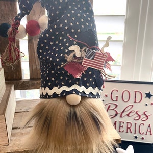 May include: A patriotic gnome with a blue and white star patterned hat, a white beard, and a red, white, and blue American flag on the hat. The gnome is sitting on a wooden surface with a sign that says "God Bless America" in the background.