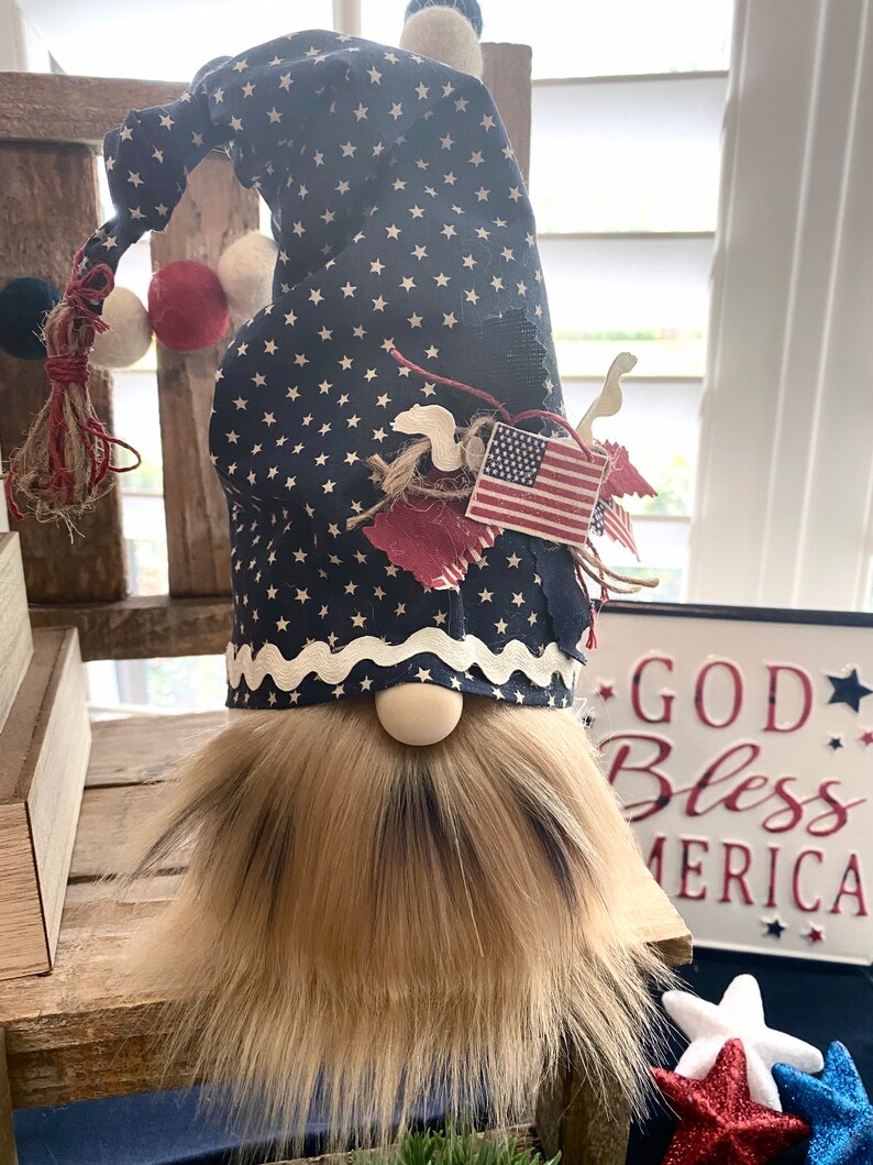 May include: A patriotic gnome with a blue and white star patterned hat, a white beard, and a red, white, and blue American flag on the hat. The gnome is sitting on a wooden surface with a sign that says "God Bless America" in the background.