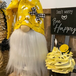 May include: A yellow gnome with a white beard and a yellow hat with black bees and a black and white bow. The gnome is sitting on a wooden bench with a sign that says "Don't worry, Bee Happy". There is a black mug with white lettering that says "SLURP" and a yellow whipped cream topping with a black bee on top.