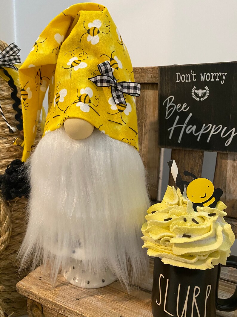 May include: A yellow gnome with a white beard and a yellow hat with black bees and a black and white bow. The gnome is sitting on a wooden bench with a sign that says "Don't worry, Bee Happy". There is a black mug with white lettering that says "SLURP" and a yellow whipped cream topping with a black bee on top.