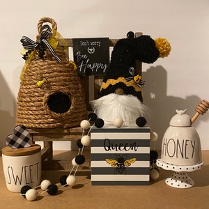May include: A black and white striped sign with the text "Queen" and a yellow bee illustration. A black and white gnome with a yellow and black hat sits on a wooden shelf with a beehive, a honey jar, and a jar labeled "Sweet". The sign reads "Don't worry, Bee Happy".