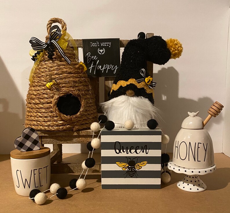 May include: A black and white striped sign with the text "Queen" and a yellow bee illustration. A black and white gnome with a yellow and black hat sits on a wooden shelf with a beehive, a honey jar, and a jar labeled "Sweet". The sign reads "Don't worry, Bee Happy".