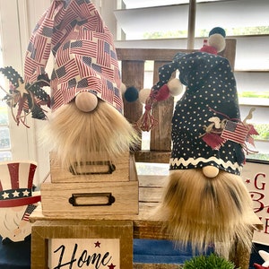 May include: Two patriotic gnomes with red, white, and blue fabric hats. The gnome on the left has a hat with an American flag pattern. The gnome on the right has a hat with blue stars on a white background. Both gnomes have brown beards and are sitting on wooden shelves. The shelves are decorated with a sign that says "Home of the Brave".