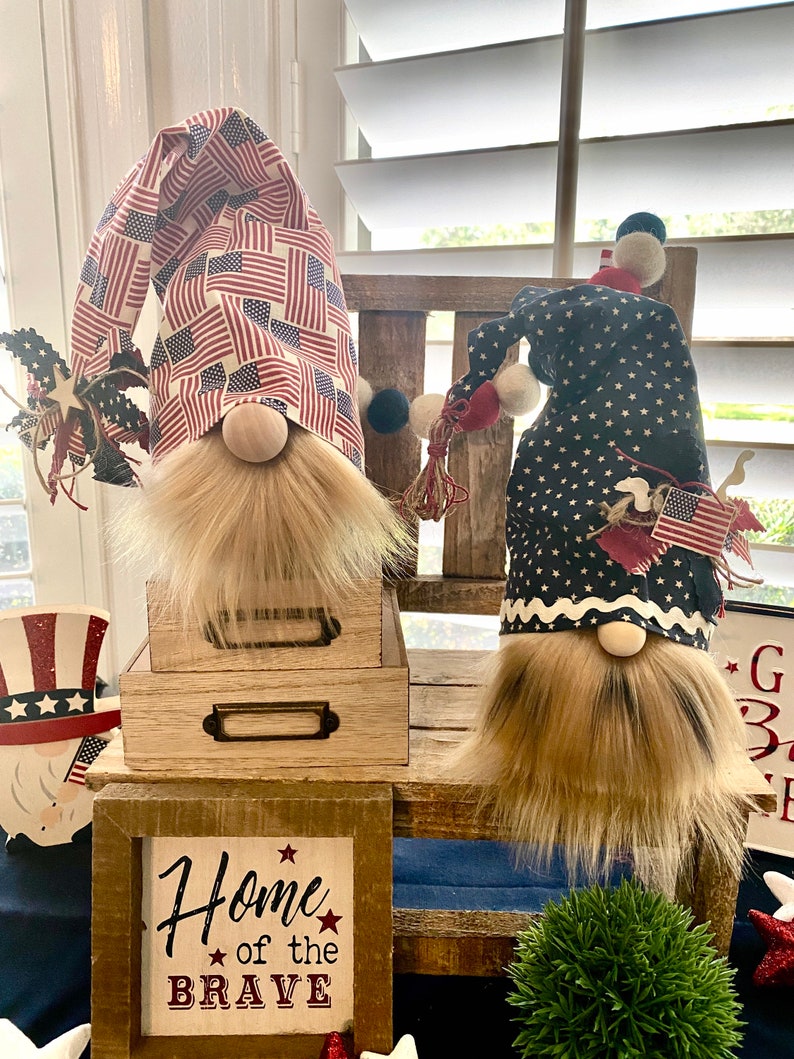 May include: Two patriotic gnomes with red, white, and blue fabric hats. The gnome on the left has a hat with an American flag pattern. The gnome on the right has a hat with blue stars on a white background. Both gnomes have brown beards and are sitting on wooden shelves. The shelves are decorated with a sign that says "Home of the Brave".