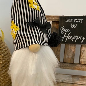 May include: A black and white striped gnome with a yellow bee design on the hat. The gnome has a white beard and is sitting on a wooden bench. A sign behind the gnome reads "Don't worry, Bee Happy".