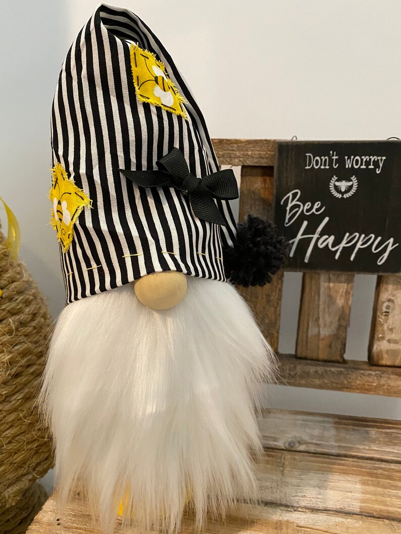May include: A black and white striped gnome with a yellow bee design on the hat. The gnome has a white beard and is sitting on a wooden bench. A sign behind the gnome reads "Don't worry, Bee Happy".