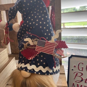 May include: Patriotic gnome with a blue and white star patterned hat, red, white, and blue fabric accents, and a long brown beard. The gnome is standing on a wooden surface with a sign that says "God Bless America".