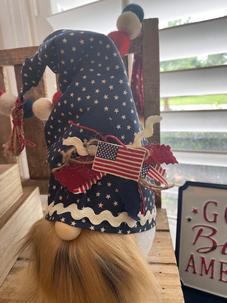 May include: Patriotic gnome with a blue and white star patterned hat, red, white, and blue fabric accents, and a long brown beard. The gnome is standing on a wooden surface with a sign that says "God Bless America".