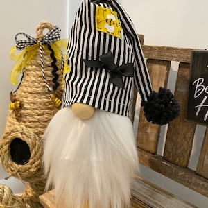 May include: A black and white striped gnome with a white beard and a black pom-pom on the top of its hat. The hat has a yellow bee patch on it and a black bow.