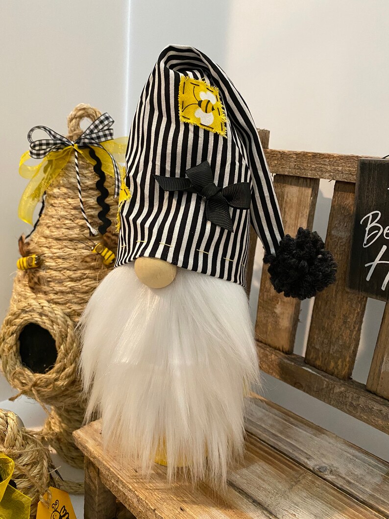 May include: A black and white striped gnome with a white beard and a black pom-pom on the top of its hat. The hat has a yellow bee patch on it and a black bow.