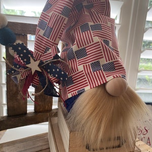 May include: A patriotic gnome with a red, white, and blue American flag patterned hat. The gnome is sitting on a wooden box with a drawer. The gnome has a long, white beard.