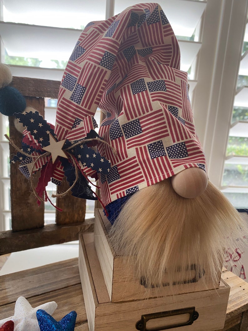 May include: A patriotic gnome with a red, white, and blue American flag patterned hat. The gnome is sitting on a wooden box with a drawer. The gnome has a long, white beard.