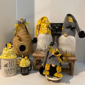 May include: Three bee-themed gnomes, one with a black and white striped hat, one with a yellow hat with bees, and one with a black hat with a yellow bee. The gnomes are sitting on a wooden stool and a small white pedestal. There are two bee-themed mugs and a beehive-shaped decoration.