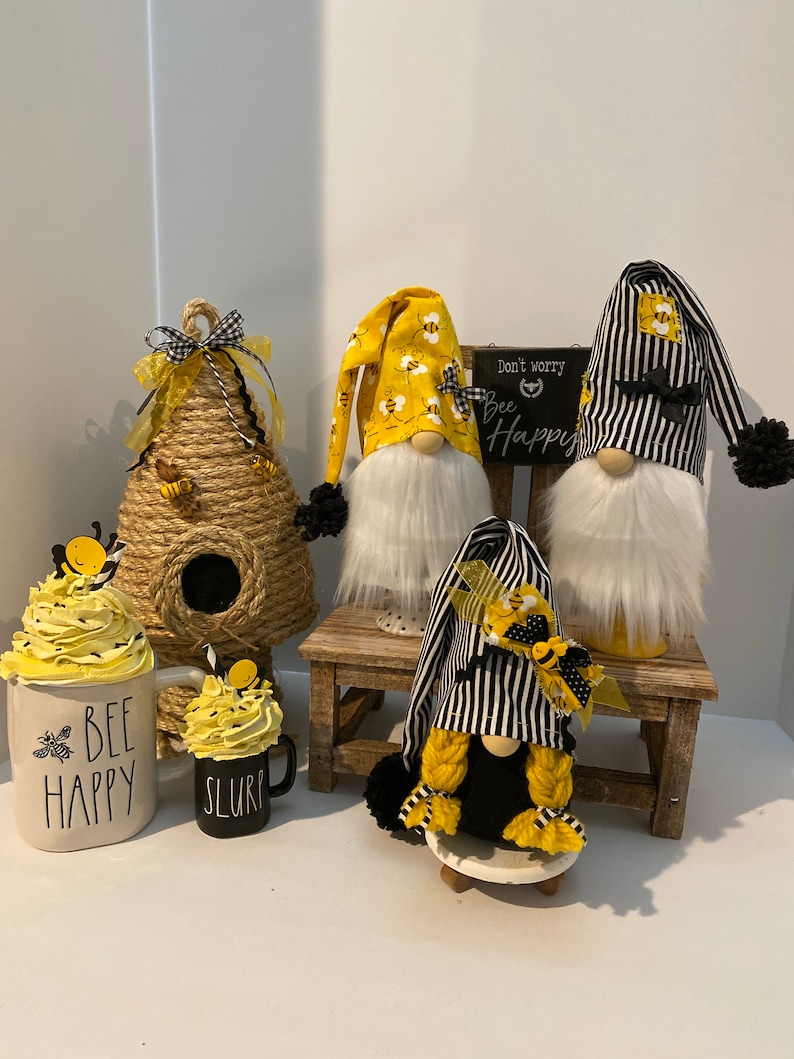 May include: Three bee-themed gnomes, one with a black and white striped hat, one with a yellow hat with bees, and one with a black hat with a yellow bee. The gnomes are sitting on a wooden stool and a small white pedestal. There are two bee-themed mugs and a beehive-shaped decoration.