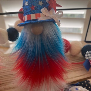 May include: Patriotic gnome decoration with a blue, white, and red glitter hat, a white bow, and a red, white, and blue fur beard.
