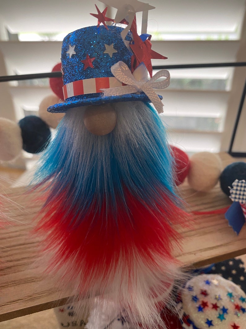 May include: Patriotic gnome decoration with a blue, white, and red glitter hat, a white bow, and a red, white, and blue fur beard.