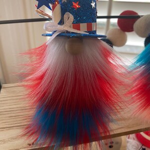 May include: A patriotic gnome decoration with a blue, white, and red hat and beard. The hat is decorated with stars and stripes and a white bow. The beard is made of fluffy red, white, and blue fur.