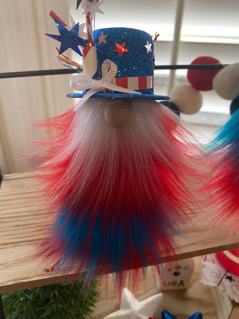 May include: A patriotic gnome decoration with a blue, white, and red hat and beard. The hat is decorated with stars and stripes and a white bow. The beard is made of fluffy red, white, and blue fur.