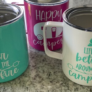 May include: Three stainless steel travel mugs with different designs. The first mug is teal green with white text that reads "Life is better around the campfire". The second mug is pink with white text that reads "Happy Camper". The third mug is white with green text that reads "Life is better around the campfire".