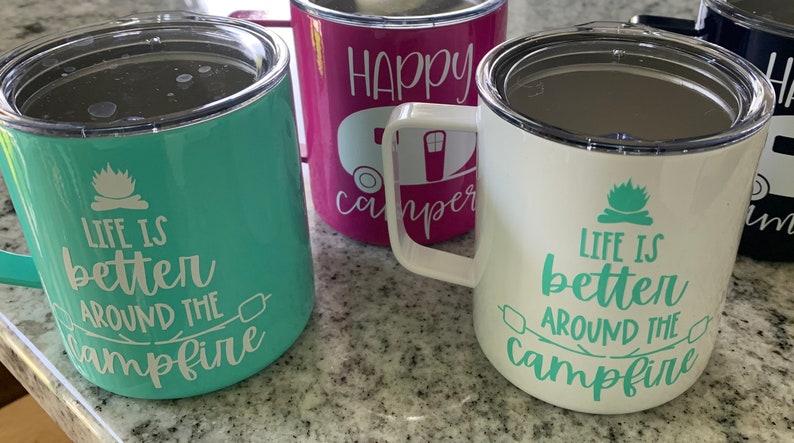 May include: Three stainless steel travel mugs with different designs. The first mug is teal green with white text that reads "Life is better around the campfire". The second mug is pink with white text that reads "Happy Camper". The third mug is white with green text that reads "Life is better around the campfire".