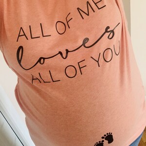 May include: A peach-coloured t-shirt with the text "All of me loves all of you" in black. The shirt also features a pair of black footprints on the lower front.