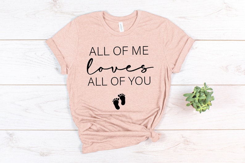 May include: A light pink t-shirt with the text "All of me loves all of you" and a pair of black footprints.