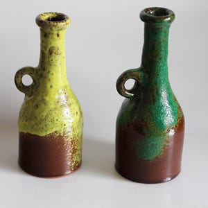 Puede incluir: Dos jarrones de cerámica con forma de botella. Uno es amarillo y marrón, el otro verde y marrón. Cada uno tiene un asa pequeña y un cuello estrecho. Los jarrones son probablemente objetos decorativos.