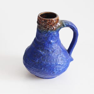 May include: A blue ceramic jug with a handle. The jug has a textured surface and a brown rim. The handle is curved and attached to the side. It is likely used for holding liquids, such as water or milk.