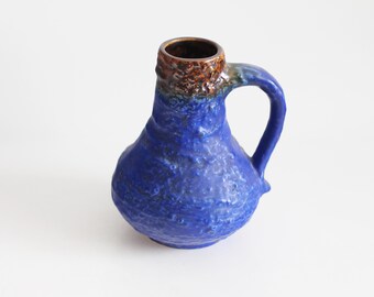 May include: A blue ceramic pitcher with a handle. The pitcher has a textured surface and a brown rim. The handle is curved and attached to the side of the pitcher. The pitcher is likely used for holding liquids.