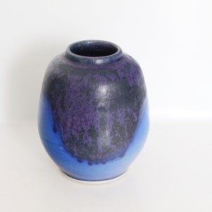 Puede incluir: Un jarrón de cerámica con forma redondeada y una abertura ancha. El jarrón presenta un degradado de colores, pasando de un azul profundo en la base a un morado oscuro y negro en la parte superior. El jarrón mide unos 20 cm de alto.