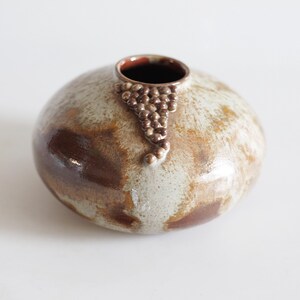 Puede incluir: Un jarrón de cerámica redondo con un esmalte moteado marrón y beige. Un grupo de pequeños puntos marrones en relieve adorna la parte delantera. El interior del jarrón es de color marrón oscuro. El jarrón tiene unos 13 cm de diámetro.