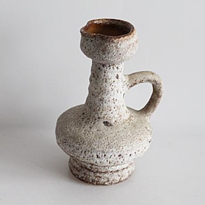 Könnte beinhalten: Eine strukturierte, cremefarbene Keramikvase mit Henkel und kleiner Öffnung. Die Vase hat einen bauchigen Boden, einen schlanken Hals und eine abgerundete Oberseite. Das Innere der Öffnung ist braun. Die Vase wird wahrscheinlich zu Dekorationszwecken verwendet.