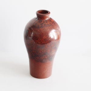Könnte beinhalten: Eine braune Keramikvase mit einem gesprenkelten Überzug. Die Vase hat einen runden Körper und einen schmalen Hals.