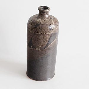 Könnte beinhalten: Eine handgefertigte Keramikflasche mit dunkler, strukturierter Oberfläche. Die Flasche hat einen schmalen Hals und einen abgerundeten Körper mit einem Farbverlauf von Dunkelbraun zu Schwarz. Die Oberfläche hat ein gesprenkeltes Aussehen, was ihren rustikalen Charme unterstreicht. Für die Inneneinrichtung geeignet.