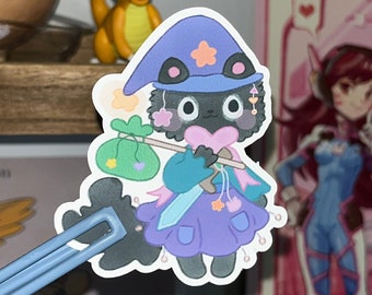 Peut inclure: Un autocollant de dessin animé noir et blanc d'un chat portant un chapeau de sorcière violet et bleu et tenant une épée et un sac de pièces d'or. Le chat a un cœur rose sur la poitrine et une étoile sur son chapeau.