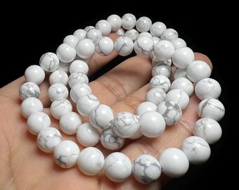 Puede incluir: Primer plano de pulseras de cuentas blancas con vetas grises. Las cuentas redondas son lisas y parecen ser de piedra. Las pulseras se sostienen en una mano sobre un fondo negro.