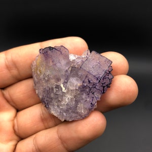 Puede incluir: Primer plano de un grupo de cristales de fluorita. El cristal es una mezcla de secciones transl&uacute;cidas moradas y claras, con una superficie rugosa y texturizada. El cristal se sostiene en una mano sobre un fondo oscuro.