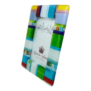 Peut inclure: Un cadre photo rectangulaire composé de segments de verre fusionné multicolores dans des tons jaune, bleu, vert, rouge et blanc. Le cadre présente des accents décoratifs millefiori et contient un insert blanc avec un dessin au trait noir de Venise et le texte Queen of glass Venezia - Murano.