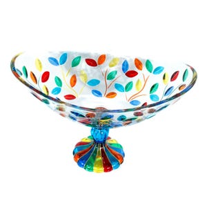 May include: A clear glass pedestal bowl featuring a hand-painted design of colourful leaves in shades of red, orange, yellow, blue, and green. The bowl has an oval shape and rests on a fluted, multi-coloured base with vertical stripes of bright colours.
