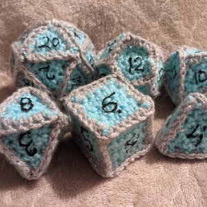 May include: A set of seven crocheted dice in various shapes, including a d20, d12, d10, d8, d6, and d4. The dice are light blue with gray trim and black numbers. The dice are handmade and suitable for tabletop gaming.