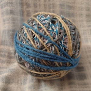May include: A ball of yarn with a mix of colors, including blue, gray, tan, and white. The yarn is tightly wound, with a prominent band of blue yarn wrapped around the outside. This ball of yarn is suitable for knitting or crocheting projects.