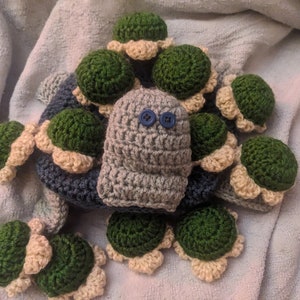 May include: A handmade crochet turtle toy with a gray body, green shell, and tan edging. The turtle has two blue button eyes. The toy is surrounded by other crochet turtle shells.