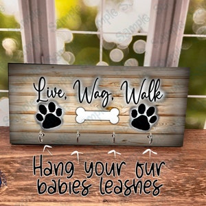 May include: A wooden sign with the text "Live, Wag, Walk" and two paw prints. The sign has four hooks for hanging leashes. The text "Hang your our babies leashes" is below the sign.