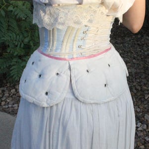 May include: A light blue and white striped corset featuring white lace trim at the top edge and white lacing at the back. Below the corset is a white quilted hip pad with silver grommets and a thin pink ribbon trim, worn over a white skirt.