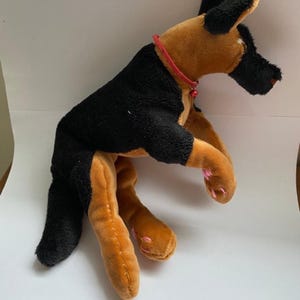 May include: A soft toy German Shepherd dog with black and tan fur, a red collar, and pink paw pads. The toy is designed to resemble a real dog, with a detailed head and body.