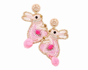 May include: A pair of pink beaded bunny earrings. The earrings feature a pink pom-pom tail, a pink gem, and a beaded design with gold accents. The earrings have a post back.