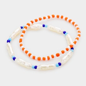May include: Two beaded bracelets. One bracelet features white, oblong beads interspersed with blue beads. The other bracelet has orange and white beads. Both bracelets are on a white background.