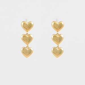 May include: A pair of gold-colored earrings, each featuring three heart-shaped charms suspended vertically. The hearts have a smooth, polished finish and are connected by delicate links. The earrings are displayed against a plain white background.
