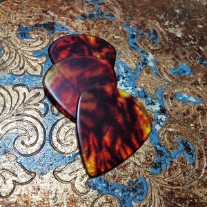 Könnte beinhalten: Drei Gitarrenplektren mit einem Schildpattmuster in Braun-, Rot- und Gelbtönen. Die Plektren sind auf einer dekorativen Oberfläche mit Gold-, Blau- und Brauntönen angeordnet.